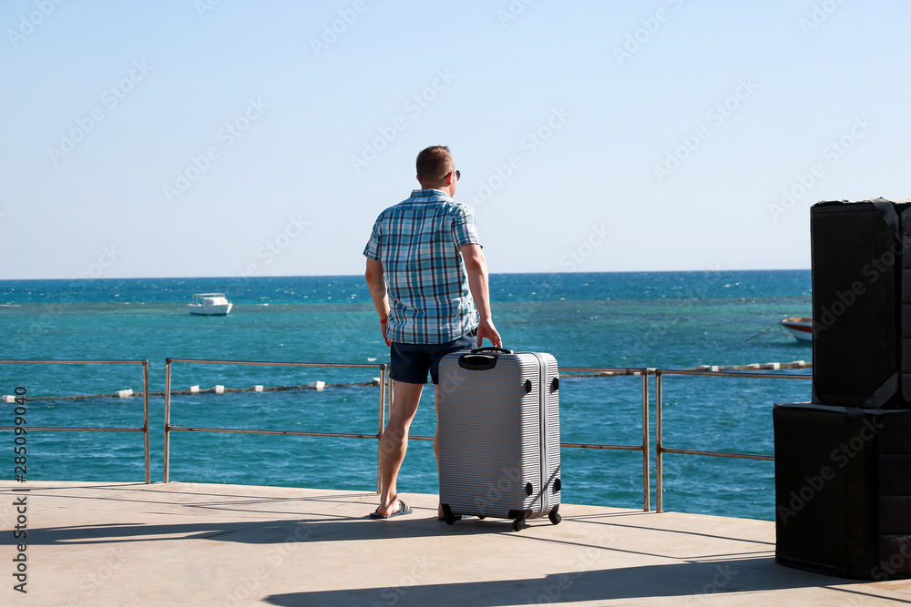 Fototapeta premium Tourist and traveler is stand on beach of hotel resort with traveled suitcase, enjoys view sea environment, nature landscape, panorama, horizon and rest in sun. Handsome man on vacation in summertime.