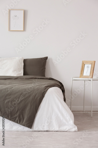 Graphic in wooden frame on white metal nightstand next to comfortable bed with black and grey pillows and duvet © Photographee.eu