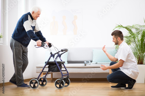 Elderly grandfather with walker trying to walk again and helpful male nurse supporting him photo