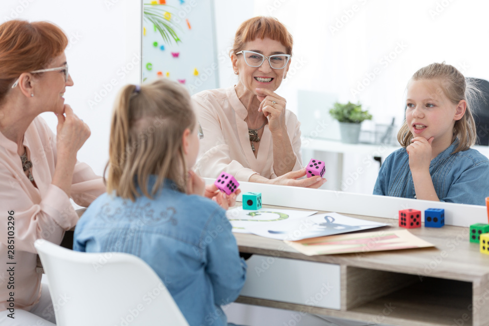 Cute blond schoolgirl looking and the mirror during speech therapy with senior doctor Cute blond schoolgirl looking and the mirror during speech therapy with senior doctor