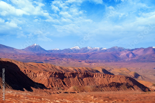 Desert landscape of red rocky formations and volcanoes near San Pedro de Atacama, Chile, against a blue sky covered by clouds.