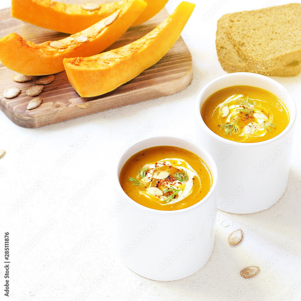 Roasted pumpkin and carrot soup with cream and pumpkin seeds on two white mug on white background with black bread slices.Vegetarian eating.Diet and healthy food concept.Space for copy and text
