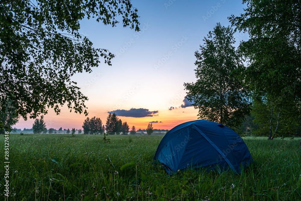 A blue tent located in a clearing in the forest. Camping tent on the background of dawn.