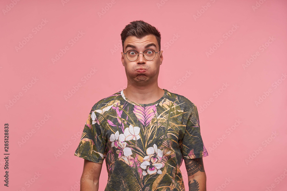 Obraz premium Indoor shot of cute guy in flowered t-shirt with wide eyes open standing over pink background, trying to hold breath, making funny grimace