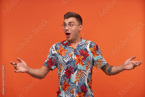 Surprised young man in floral shirt shrugs with opened mouth, standing over orange background, wearing glasses, looking away