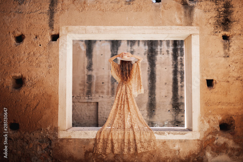 Back view of gorgeous woman in beige lace dress standing on wall of deserted building and holding hat photo