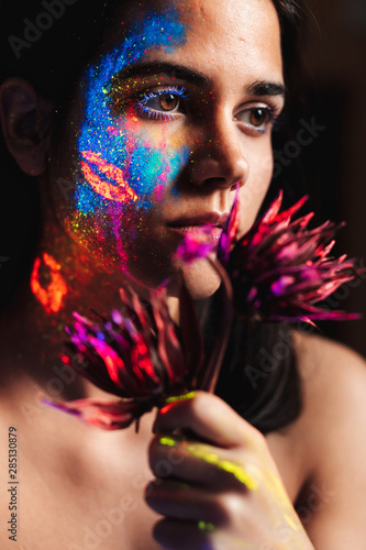 Portrait of beautiful young woman covered with luminous paint on face holding a flower and looking away. photo