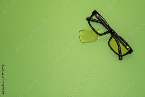 Broken glasses lie on a green surface. Nearby lies a yellow lens that has fallen out of the frame. View from above