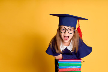Cheerful schoolgirl in graduation outfit smiling while leaning on pile of colourful books over yellow background