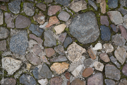 Full frame image of multicolored cobblestone with ground and green moss between stones. Top view of the old paved sidewalk. High resolution background or texture