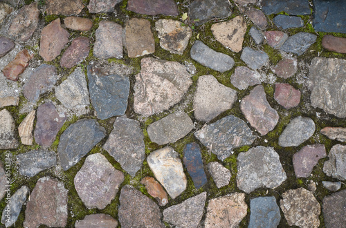 Full frame image of multicolored granite cobblestone with green moss between stones. Top view of the urban paved sidewalk. High resolution texture or background