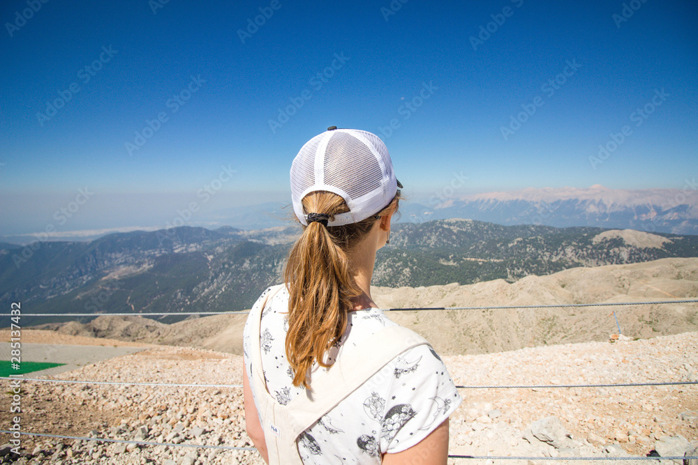 Beautiful and young girl looks at the mountains. She stands on top and looks down. A beautiful panoramic view of the mountains opens in front of her.