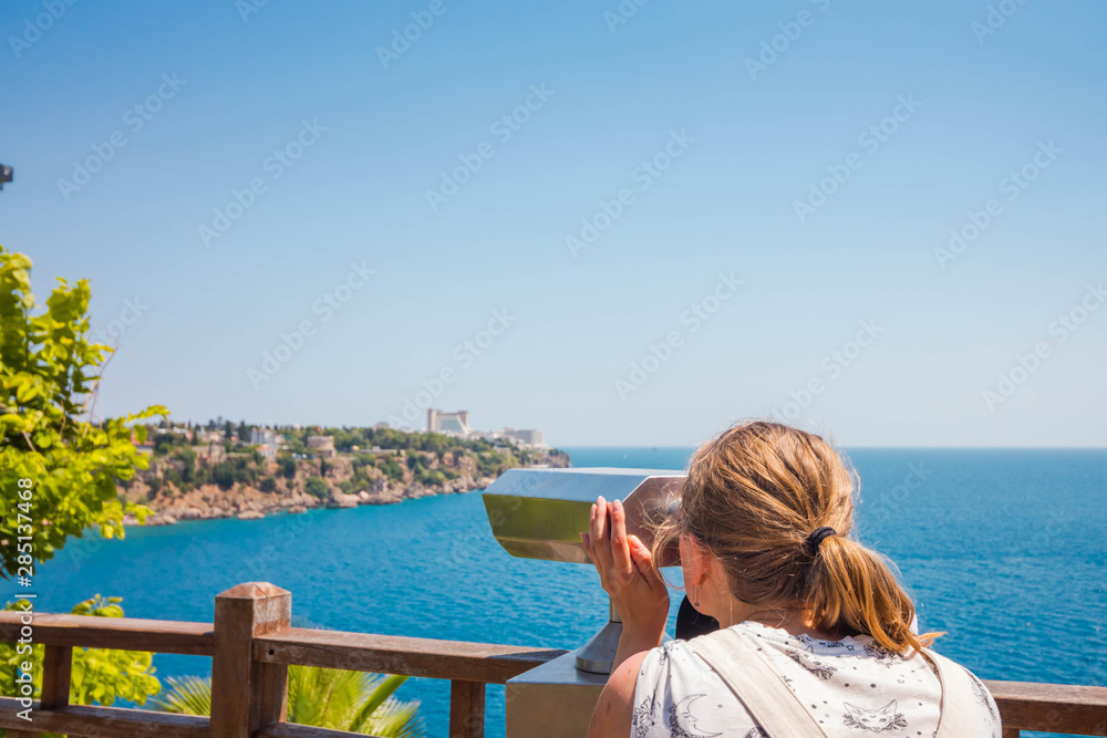 Beautiful and young girl looking through binoculars. A girl looks at a city that is far from her. The city is built on a cliff that rises above the clear sea.