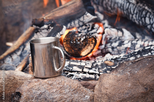Stainless mug of hot drink standing on a stone by an outdoor campfire. Travel background. Campsite, picnic in wild forest. Bonfire in camp, adventure scene