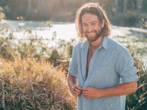 Young cheerful bearded hipster man unbuttoning blue shirt preparing to go swimming in river on sunny summer day photo