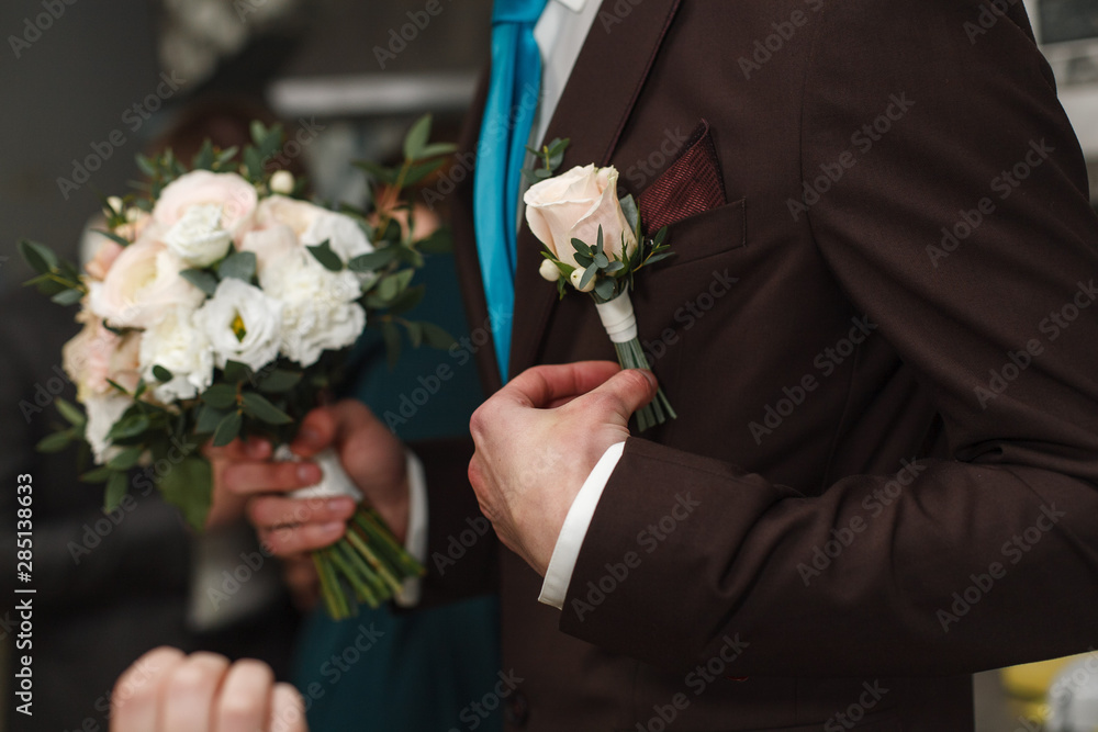 the man corrects the boutonniere the groom with a boutonniere meeting of the groom.weedding morning.The groom in a white shirt and dark brown suit straightens buttonhole. the man corrects the boutonniere the groom with a boutonniere meeting of the groom.weedding morning.The groom in a white shirt and dark brown suit straightens buttonhole.