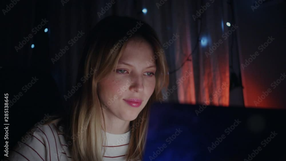 Close up beautiful blonde girl sitting at a laptop in the night and prints a message. She straightens her hair and smiles. Close up beautiful blonde girl sitting at a laptop in the night and prints a message. She straightens her hair and smiles.