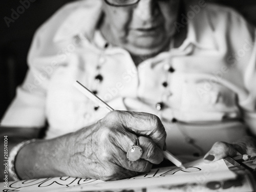 senior gray haired female in white shirt and glasses drawing on paper with pencil sitting on armchair at home photo