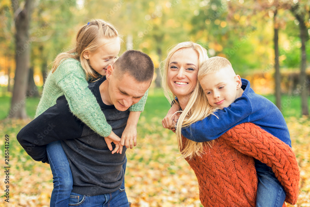 Mother and father carrying their two adorable children on shoulders look at each other during warm autumn weekend. Smiling family in autumn park. Mother and father carrying their two adorable children on shoulders look at each other during warm autumn weekend. Smiling family in autumn park.