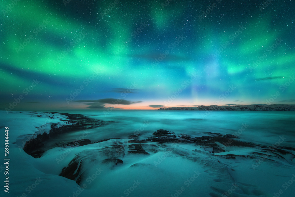 Aurora borealis over rocky beach and ocean. Northern lights in Teriberka, Russia. Starry sky with polar lights. Night winter landscape with aurora, sea with stones in blurred water, snowy mountains