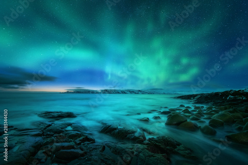 Aurora borealis over rocky beach and ocean. Northern lights in Teriberka, Russia. Starry sky with polar lights. Night winter landscape with aurora, sea with stones in blurred water, snowy mountains