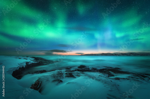 Aurora borealis over rocky beach and ocean. Northern lights in Teriberka, Russia. Starry sky with polar lights. Night winter landscape with aurora, sea with stones in blurred water, snowy mountains