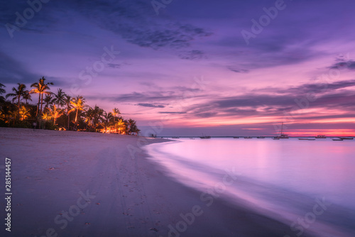 Fototapeta Naklejka Na Ścianę i Meble -  Sandy beach with palm trees at colorful sunset in summer.  Tropical landscape with sea shore, blurred water, palms, boats and yachts in ocean, purple sky with clouds at night. Travel in exotic Africa