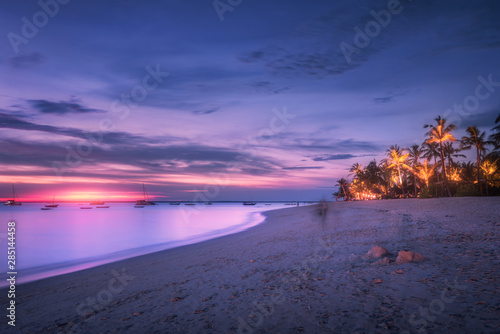 Sandy beach with palm trees at colorful sunset in summer. Tropical landscape with sea shore, blurred water, palms, boats and yachts in ocean, purple sky with clouds at night. Travel in exotic Africa