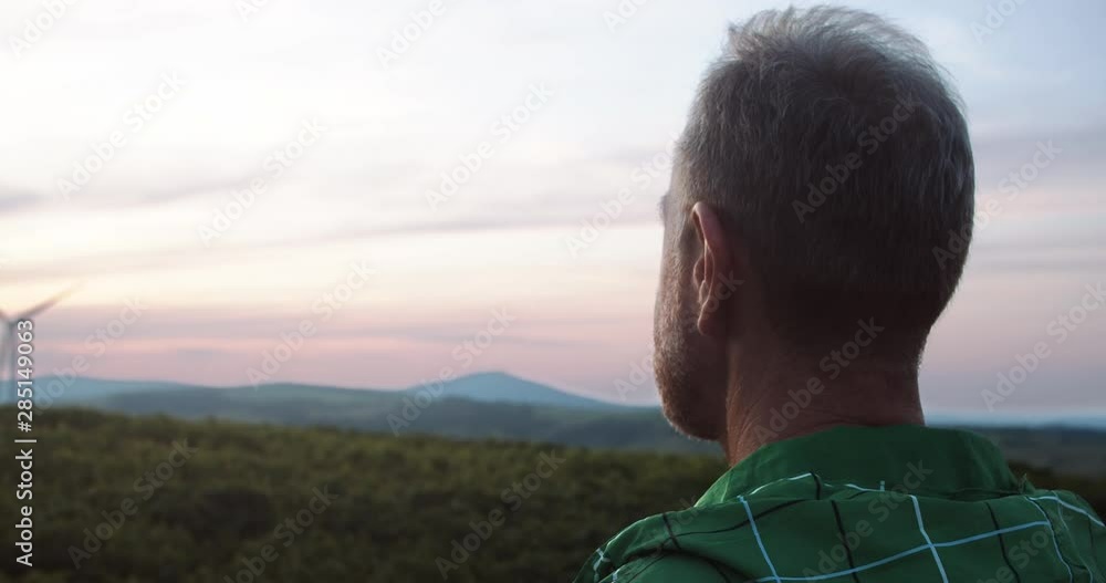 Close-up farmer watching glorious nature scenery in rural greenland with wind turbines rotating. Windpower electricity production. Eco-friendly farming concept. Close-up farmer watching glorious nature scenery in rural greenland with wind turbines rotating. Windpower electricity production. Eco-friendly farming concept.