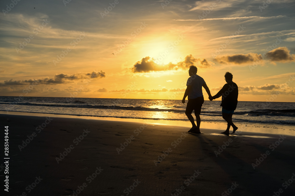 Middle aged couple holding hands walking on beach at sunrise.