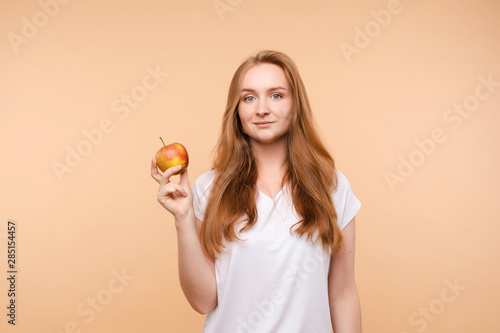 Beautiful young girl with tied on back hair eating tasty apple on lunch. Side view of attractive model promoting healthy lifestyle. Brunette woman with white teeth holding delicious fruit in her hand.