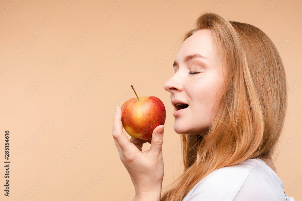 Beautiful young girl with tied on back hair eating tasty apple on lunch. Side view of attractive model promoting healthy lifestyle. Brunette woman with white teeth holding delicious fruit in her hand. Beautiful young girl with tied on back hair eating tasty apple on lunch. Side view of attractive model promoting healthy lifestyle. Brunette woman with white teeth holding delicious fruit in her hand.