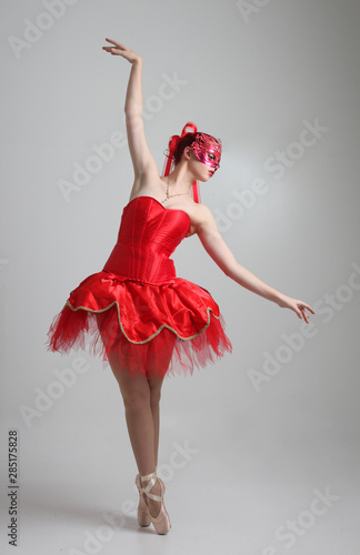 full length portrait of girl wearing red ballerina tutu and mask. dancing pose against a studio background.