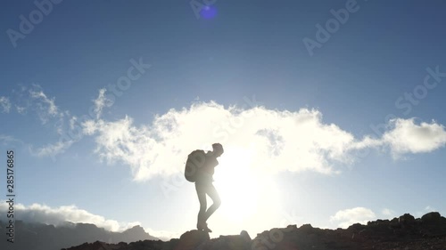 Silhouette of a young woman hiker with backpack walking uphill towards the summit against blue sky and clouds at sunset. Slow motion. Lady is hiking in beautiful mountains on Canary Islands.
