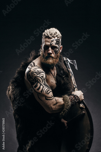 Medieval warrior berserk Viking with tattoo on skin, red beard and braids in hair with axe and shield attacks enemy. Concept historical photo