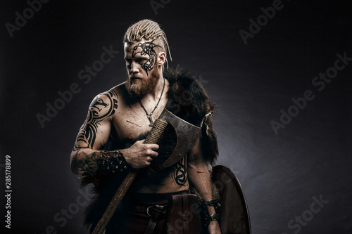 Medieval warrior berserk Viking with tattoo on skin, red beard and braids in hair with axe and shield attacks enemy. Concept historical photo photo