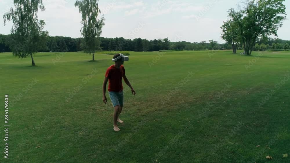 A man using a VR headset and playing a virtual game in the park. Young guy in helmet experiencing augmented reality on the background of nature. A human on the right in the frame. 4K drone footage. A man using a VR headset and playing a virtual game in the park. Young guy in helmet experiencing augmented reality on the background of nature. A human on the right in the frame. 4K drone footage.