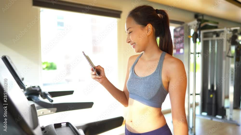A fit, beautiful young Asian women on a treadmill smiles as she uses her smartphone to respond to your text. A fit, beautiful young Asian women on a treadmill smiles as she uses her smartphone to respond to your text.