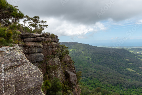A rocky and grey cliff edge with trees and vegetation on top, overlooks the dense Australia forest  on a partially cloudy day at Drawing Room Rocks in NSW, Australia