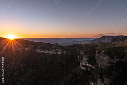 The sun starts to dip over the horizon casting a beautiful orange to blue gradient across the sky with the rocky cliffs covered in vegetation in the foreground