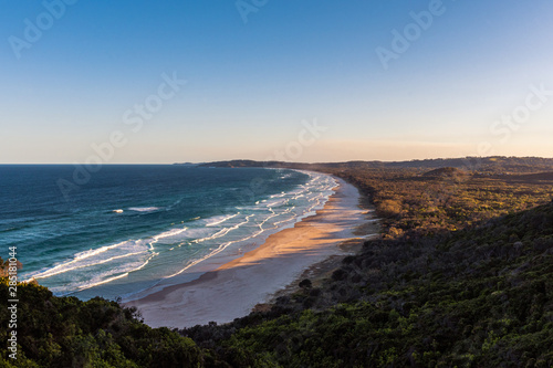The sun lowers over the lush dense forests that end at Tallow beach with the waves of the ocean rushing in. Byron Bay in NSW, Australia
