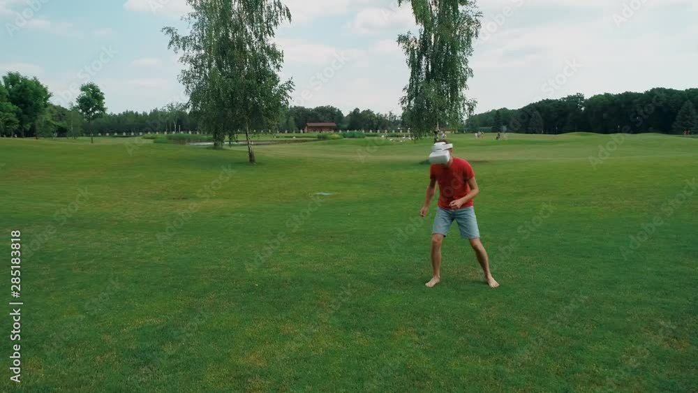 A man using a VR headset and playing a virtual game in the park. Young guy in helmet experiencing augmented reality on the background of nature. A human on the right in the frame. 4K drone footage. A man using a VR headset and playing a virtual game in the park. Young guy in helmet experiencing augmented reality on the background of nature. A human on the right in the frame. 4K drone footage.