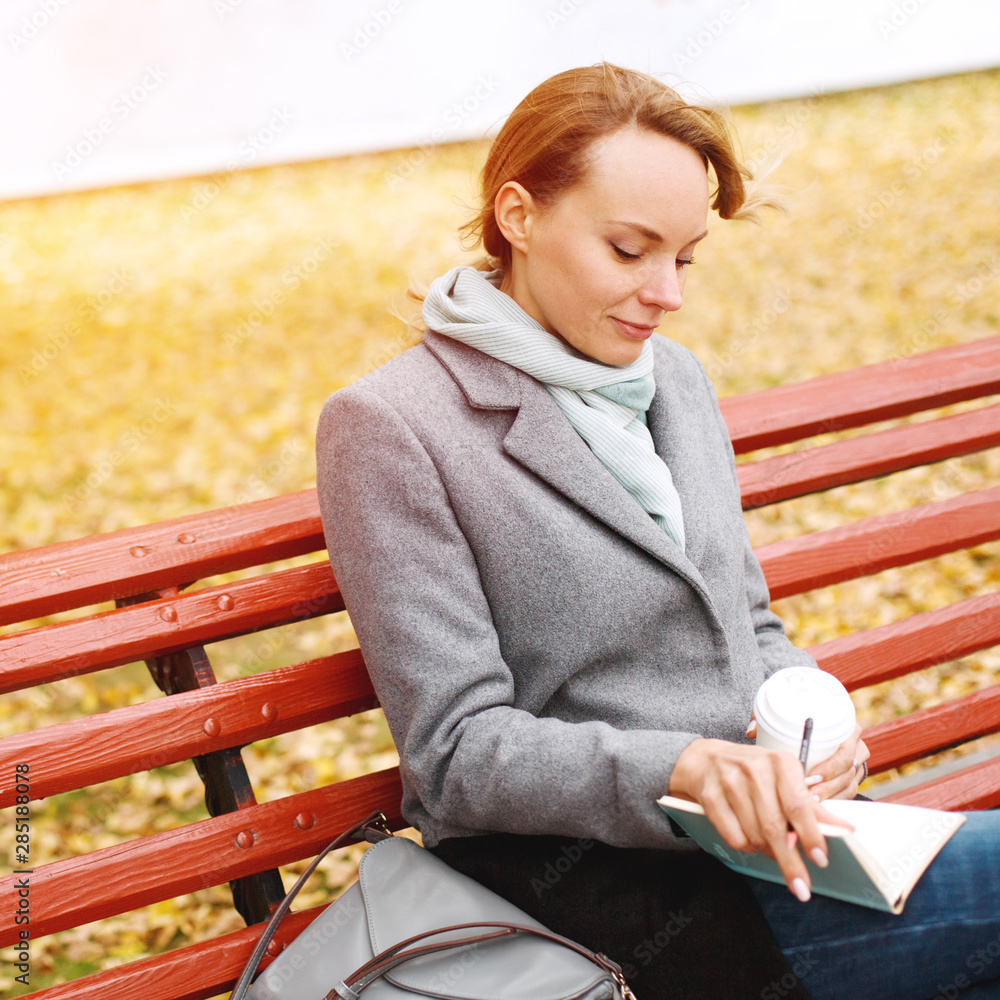 Young pretty woman sitting on a bench in autumn park with a notebook and coffee