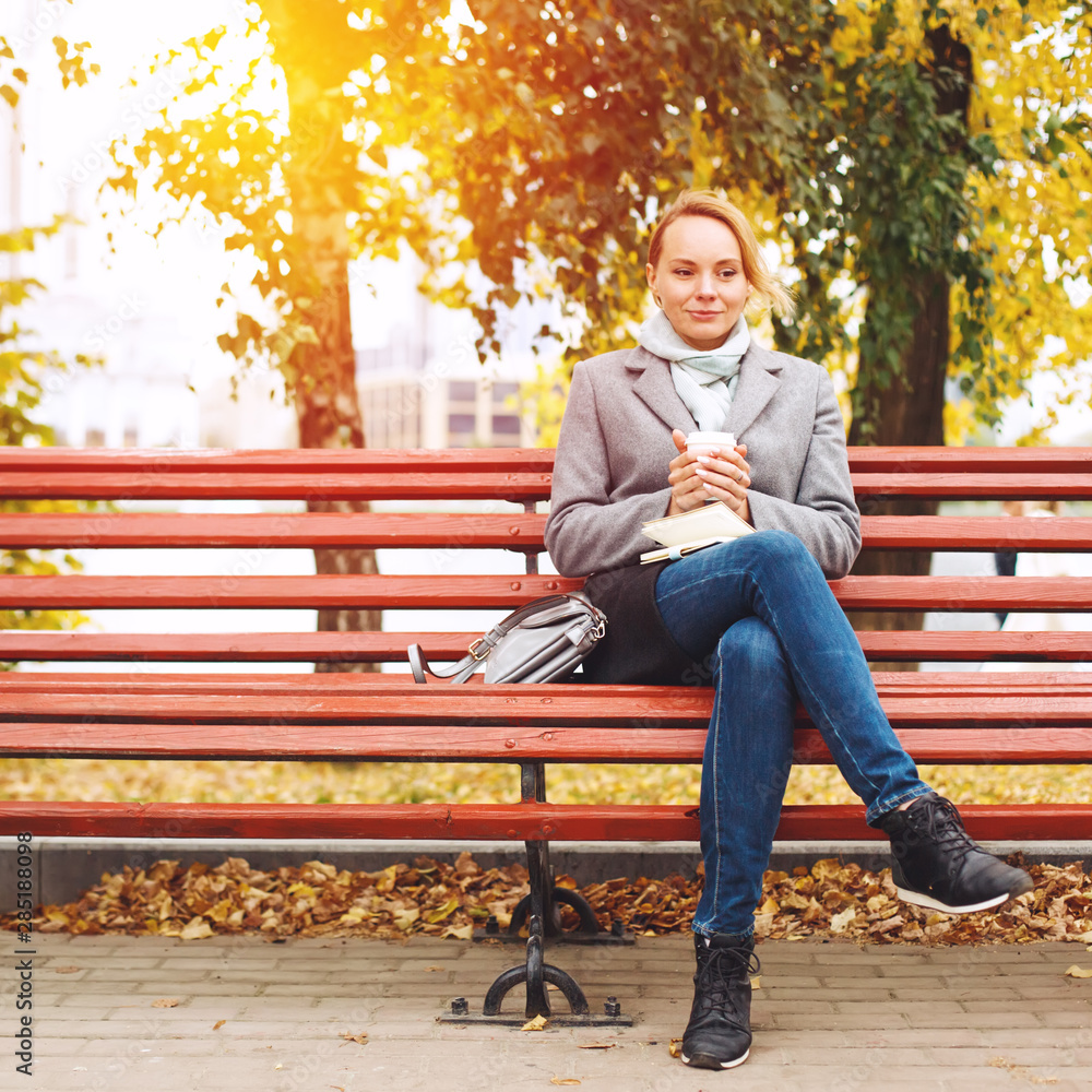 Stylish woman sitting on a bench with coffee and paper notebook, autumn sunny day
