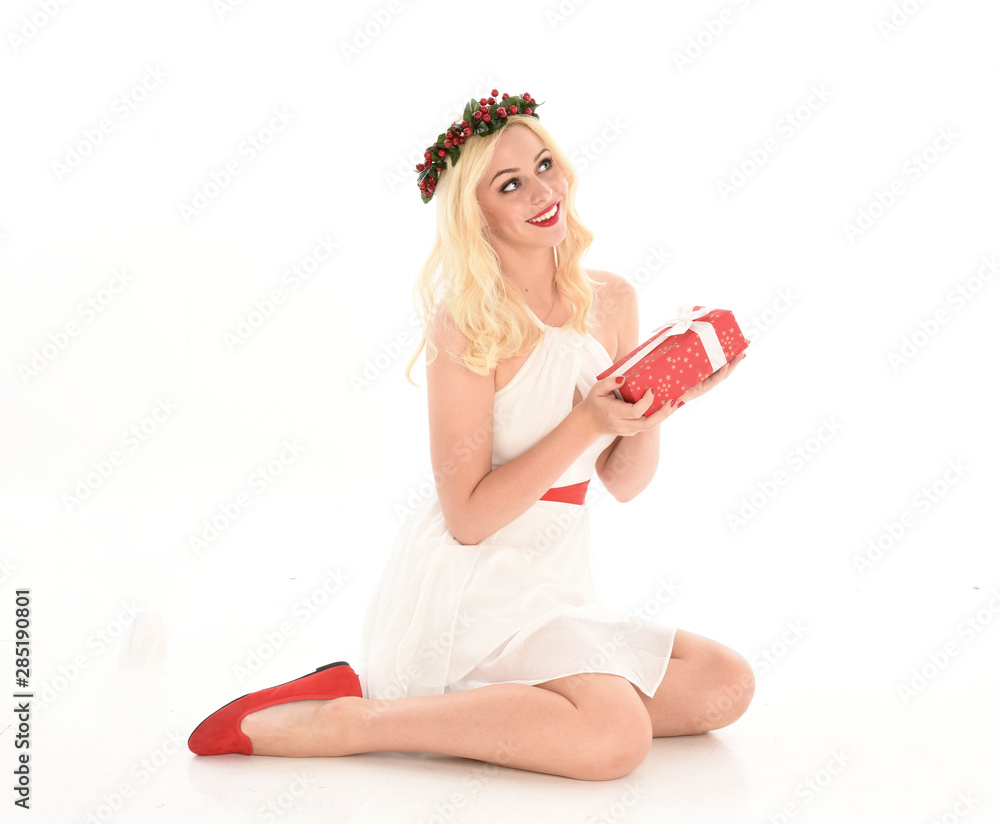 full length portrait of blonde girl wearing a white dress and flower crown. Sitting pose, holding red present, isolated against a white studio background. full length portrait of blonde girl wearing a white dress and flower crown. Sitting pose, holding red present, isolated against a white studio background.