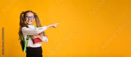 Adorable Schoolgirl Pointing Finger At Copyspace Holding Book, Yellow Background