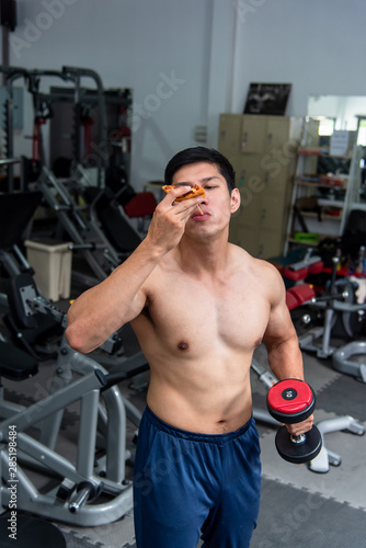 Asian strong man who loves exercising is holding a dumbbell and is eating pizza for getting energy before workout at fitness center