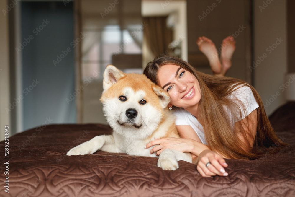 Woman lying with her akita inu dog on sofa in bedrroom, looking at the camera together.Young Woman and her dog in home Woman lying with her akita inu dog on sofa in bedrroom, looking at the camera together.Young Woman and her dog in home