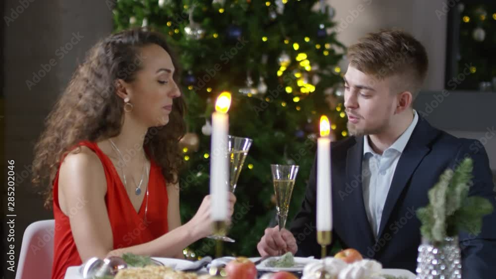 Chest-up shot of affectionate young Caucasian couple having romantic dinner date in restaurant next to Christmas tree, holding champagne flutes, wishing each other happy New Year and clinking glasses Chest-up shot of affectionate young Caucasian couple having romantic dinner date in restaurant next to Christmas tree, holding champagne flutes, wishing each other happy New Year and clinking glasses