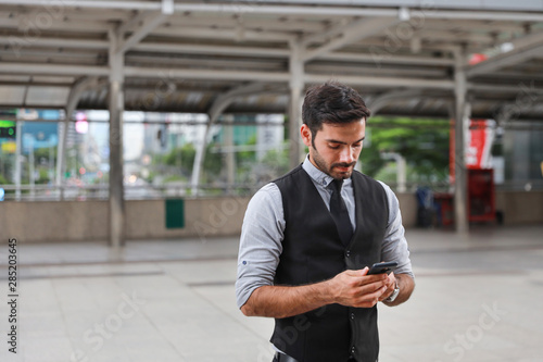 successful caucasian businessman in black suite who standing in downtown, he using cell phone while standing on walk way with city and skytrain background